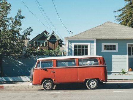 A vintage red van is parked on the street in front of a light blue house, with trees and other houses in the background.