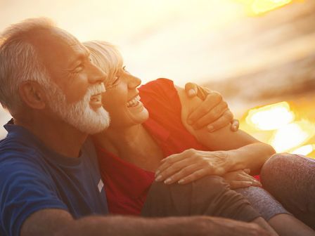 A senior couple is sitting and laughing together on a beach at sunset, enjoying the warm light and each other's company.