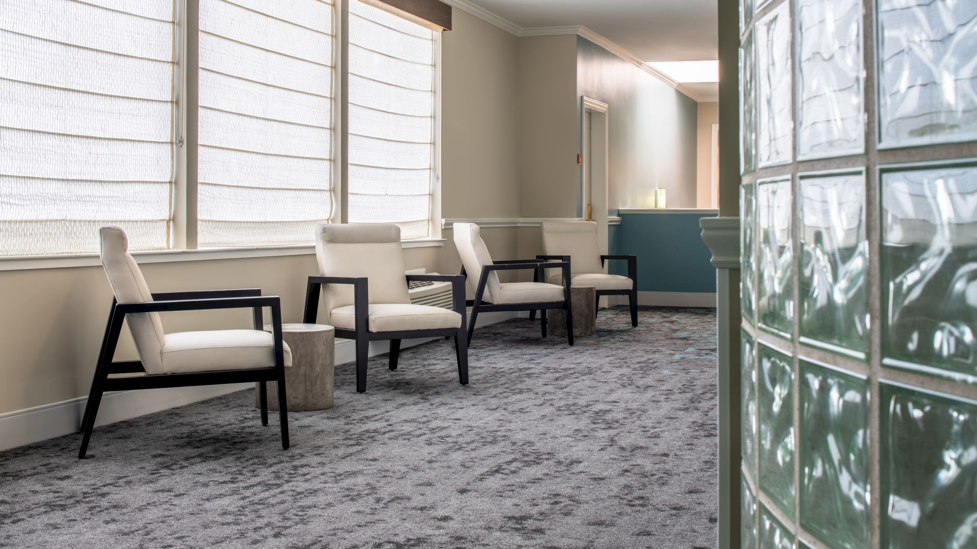 The image shows a waiting area with four chairs along a wall with large windows and sheer curtains. A reception desk is visible in the background.
