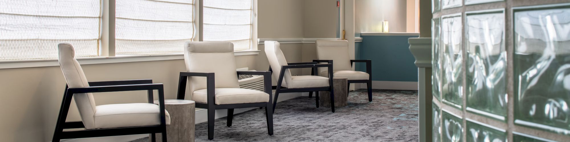 The image shows a waiting area with four chairs along a wall with large windows and sheer curtains. A reception desk is visible in the background.