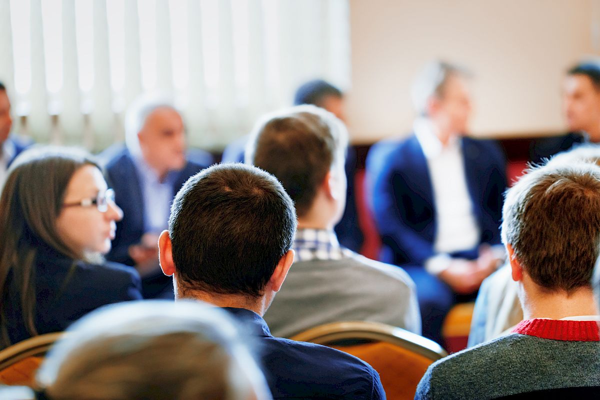 A group of people is seated in a room, engaged in a conversation or meeting. Most are looking towards the front, suggesting a presentation or discussion.
