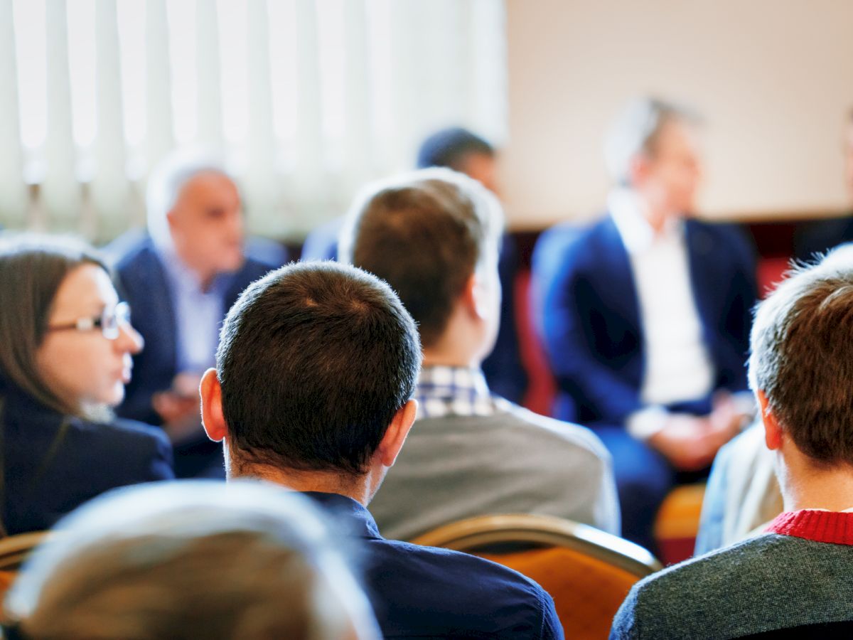 A group of people is seated in a room, engaged in a conversation or meeting. Most are looking towards the front, suggesting a presentation or discussion.