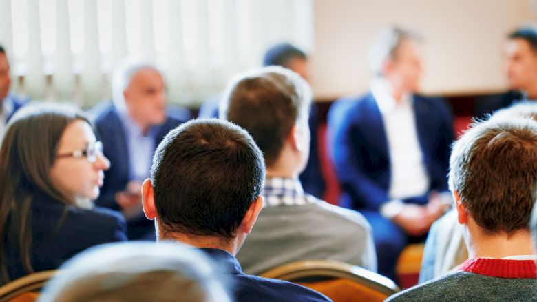 A group of people is seated in a room, engaged in a conversation or meeting. Most are looking towards the front, suggesting a presentation or discussion.