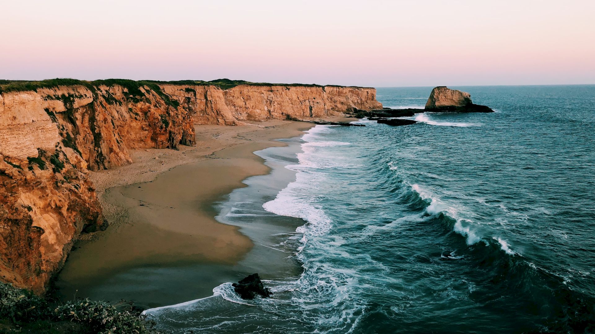 A picturesque coastline features rugged cliffs, a sandy beach, and waves crashing against the shore under a serene sunset sky.
