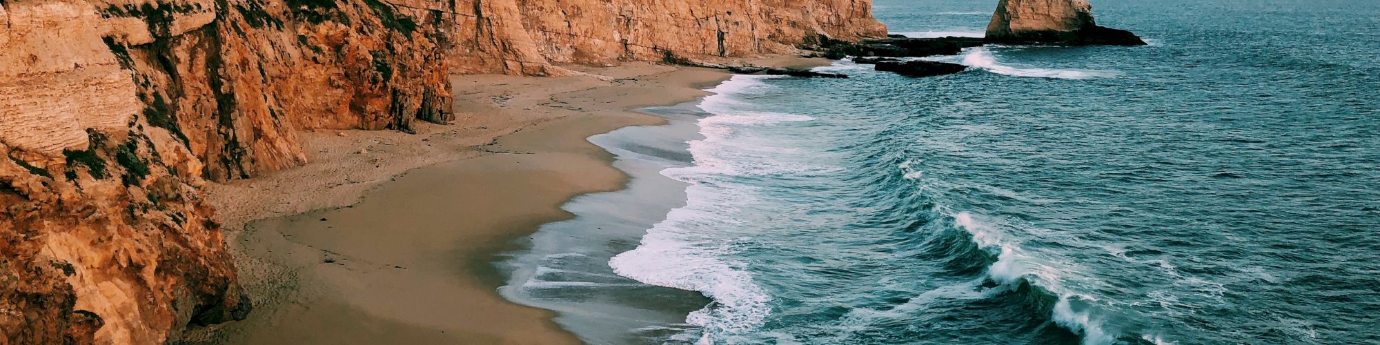 A picturesque coastline features rugged cliffs, a sandy beach, and waves crashing against the shore under a serene sunset sky.