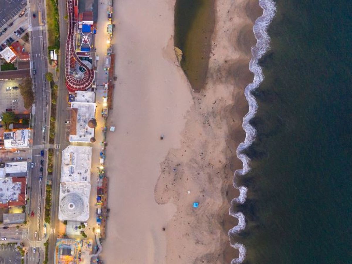 Aerial view of a beach with adjacent buildings, a roller coaster, a Ferris wheel, and waves crashing on the shore along the coastline.