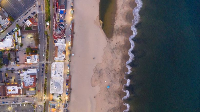 Aerial view of a beach with adjacent buildings, a roller coaster, a Ferris wheel, and waves crashing on the shore along the coastline.