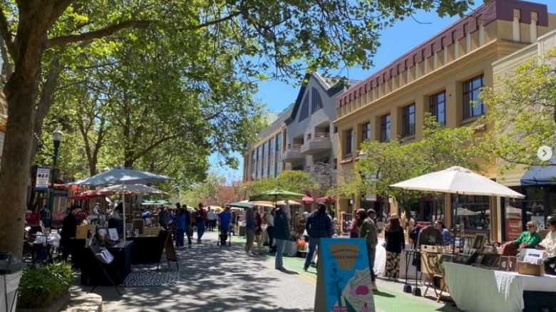 The image shows a bustling outdoor market with vendors, shoppers, trees, and buildings lining the street on a sunny day.