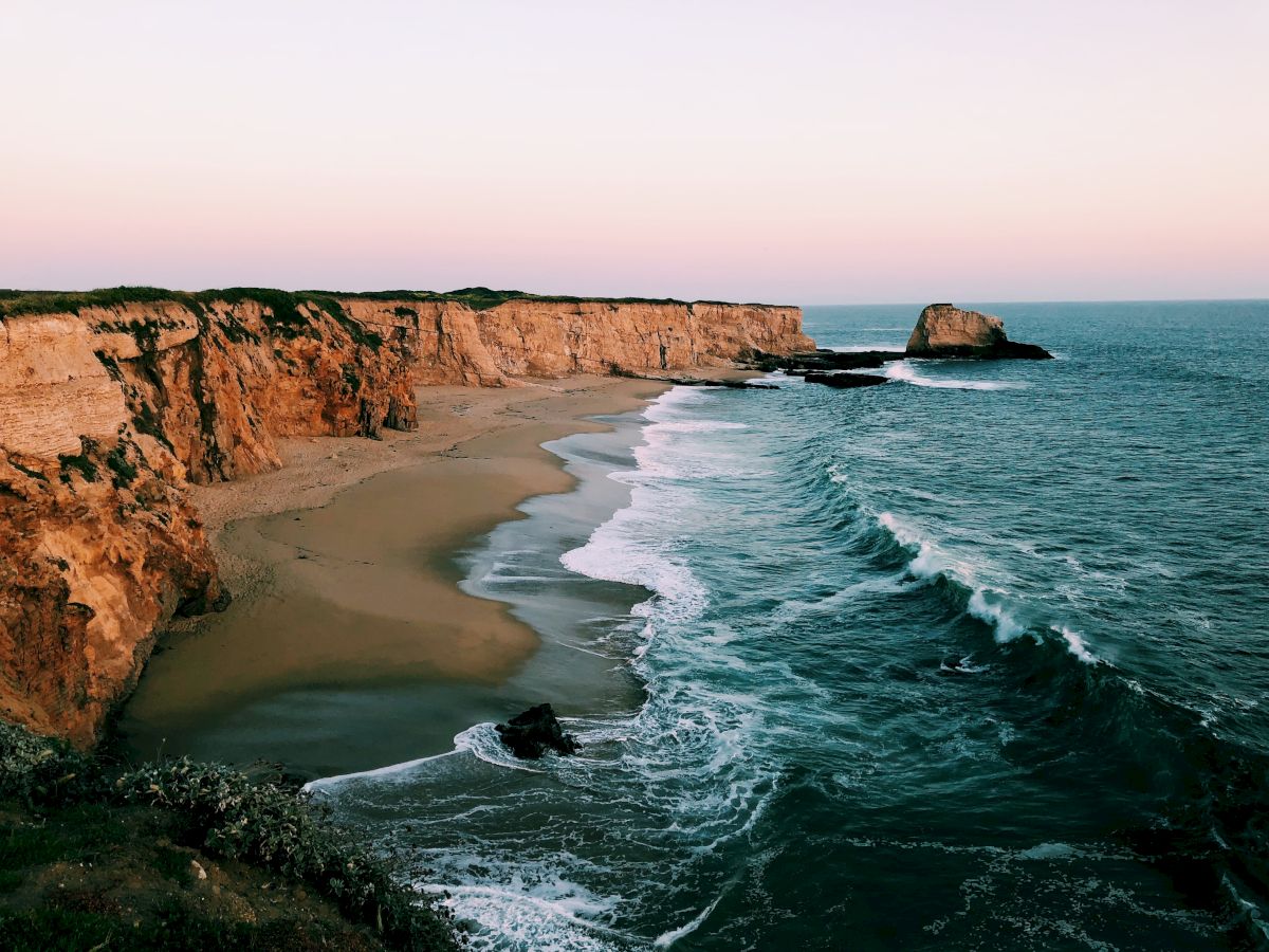 A serene coastal scene featuring tall cliffs, a sandy beach, and waves crashing against the shore under a clear sky.