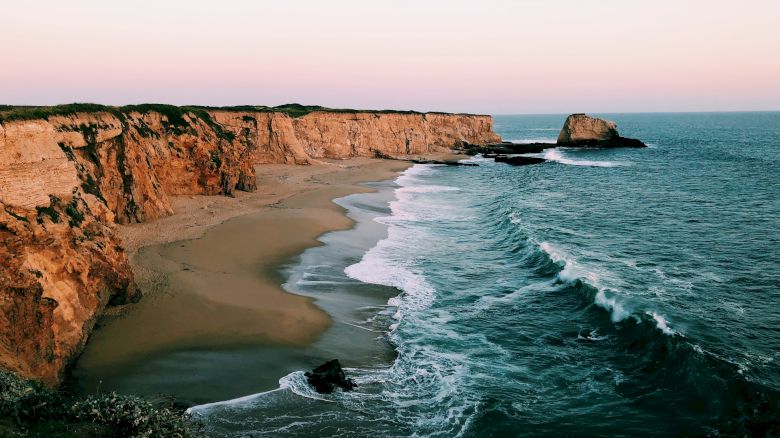 A serene coastal scene featuring tall cliffs, a sandy beach, and waves crashing against the shore under a clear sky.