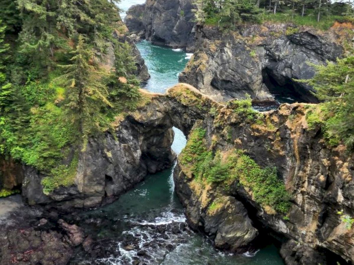 The image shows a natural rock arch over a turquoise waterway, surrounded by green vegetation and rugged cliffs, creating a scenic coastal landscape.