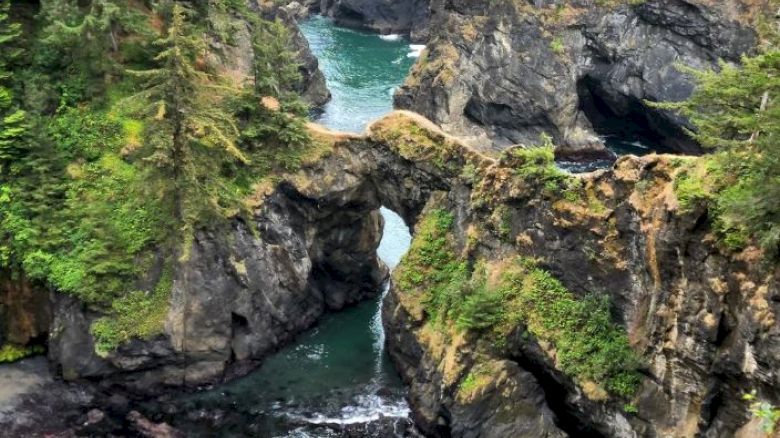 The image shows a natural rock arch over a turquoise waterway, surrounded by green vegetation and rugged cliffs, creating a scenic coastal landscape.