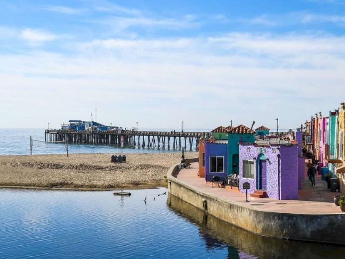 Colorful buildings line a waterfront, with a sandy beach and pier visible in the background under a blue sky with clouds.