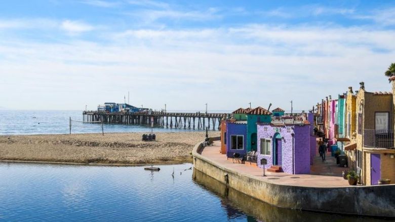 Colorful buildings line a waterfront, with a sandy beach and pier visible in the background under a blue sky with clouds.