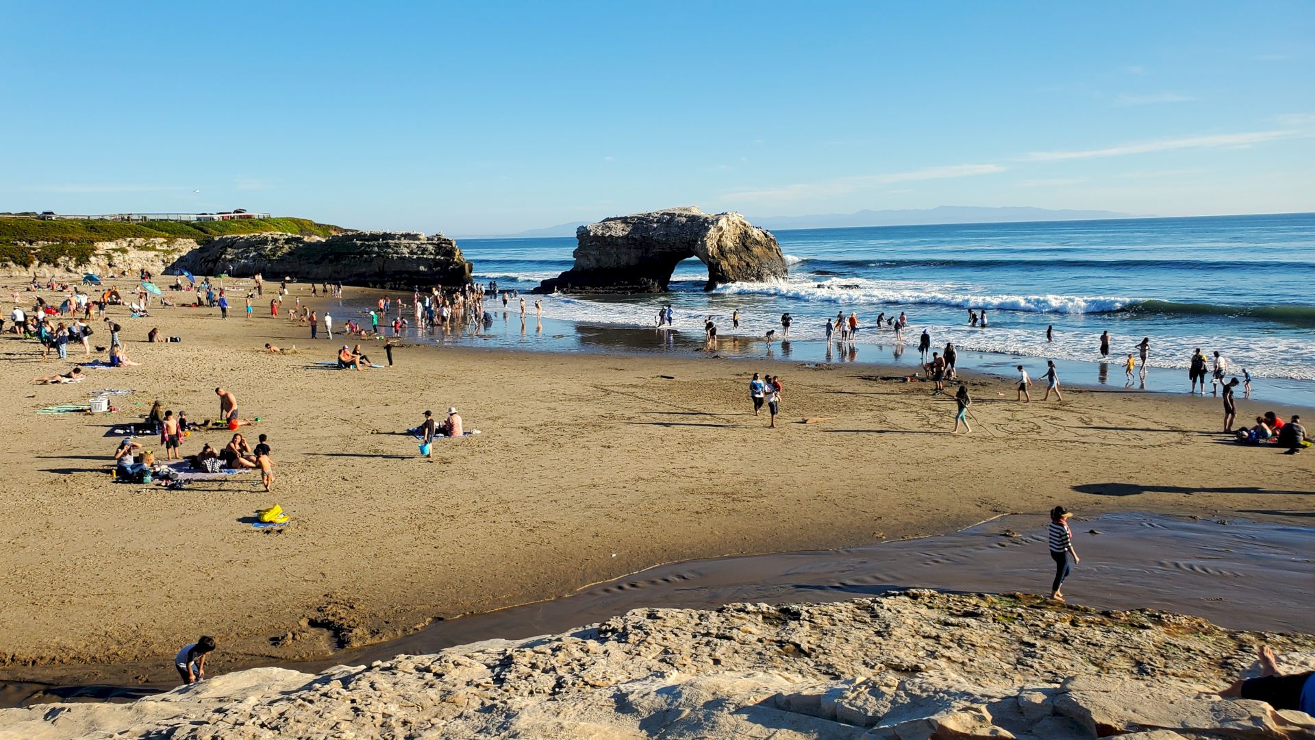 A busy beach scene with people sunbathing, wading in the water, and enjoying the day. A natural rock arch is visible in the background.