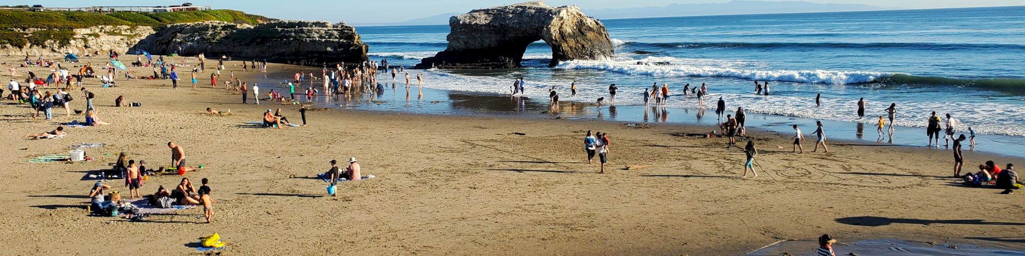 A busy beach scene with people sunbathing, wading in the water, and enjoying the day. A natural rock arch is visible in the background.