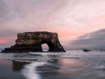 An ocean coastal landscape with a natural rock formation arch under a pink-hued sky at sunset, surrounded by waves and a serene beach.