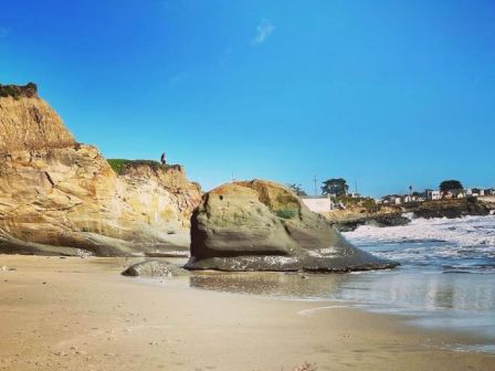 A serene beach scene features a sandy shore, a large rock formation, cliffside, and the ocean waves under a clear blue sky.