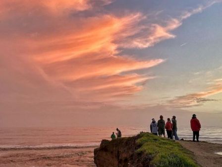 A group of people stands on a grassy cliff overlooking the ocean at sunset, with beautiful orange and pink hues in the sky, enjoying the view.