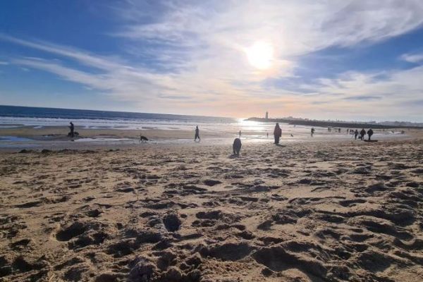 A sunny beach scene with people walking along the shore, a clear sky, and a distant lighthouse under the bright sun.