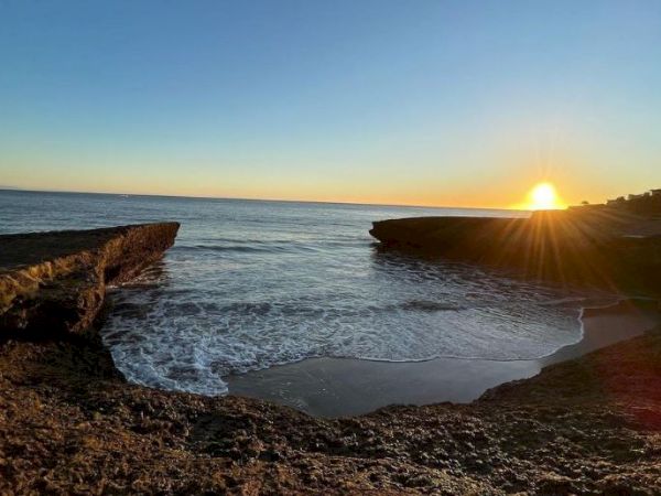 The image shows a beautiful coastal scene at sunset with rocky cliffs framing the water and the sun setting on the horizon, casting a golden glow.