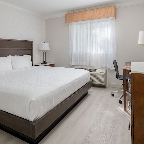 A neatly arranged hotel room with a large bed, bedside lamps, a desk with a chair, and a TV facing the bed. The window has white curtains.