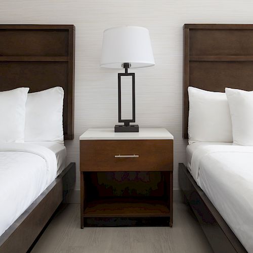 A hotel room with two neatly made beds, a wooden nightstand in the center, and a modern table lamp placed on the nightstand between the beds.