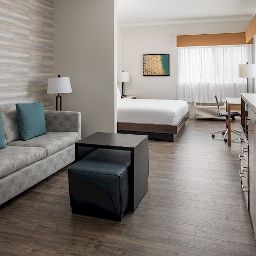 This image shows a modern hotel room with a sofa, coffee table, TV, and a bed. There's a desk near the window and two lamps in the room.