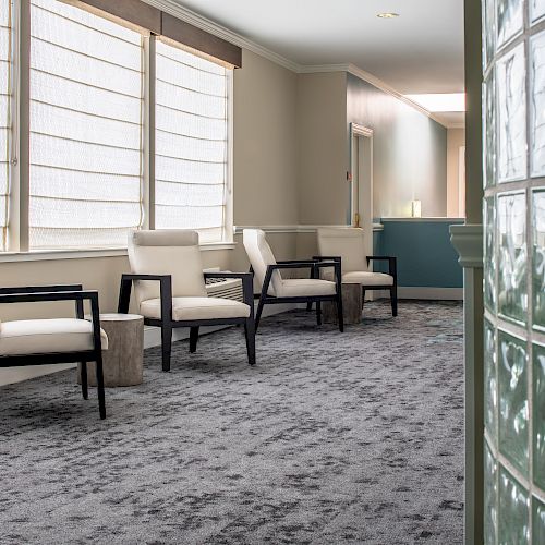 The image shows a waiting area with four modern chairs against a wall with large windows, featuring a carpeted floor and a glass block partition wall ending the sentence.