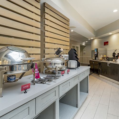 A buffet area with chafing dishes, utensils, and various equipment on a counter, adjacent to a beverage and snack station.