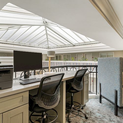 The image shows an office space with two chairs, a computer, and a printer on a desk, beside a cozy seating area, under a skylight.