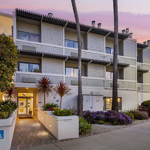 This image shows a multi-story residential building with a landscaped entrance, palm trees, and a sunset sky in the background.