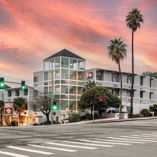 The image shows a multi-story modern building with palm trees, set against a colorful sky at dusk, and a traffic light at the intersection.