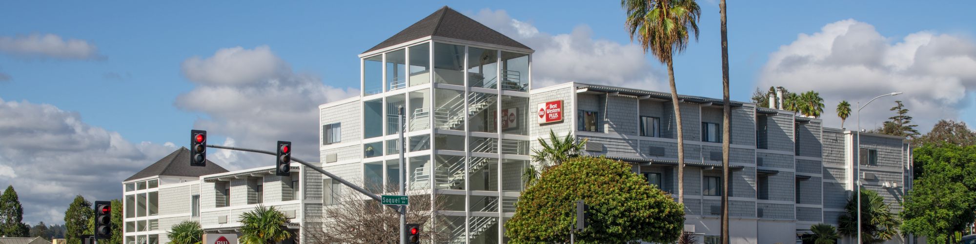 A modern white building with glass staircases, palm trees, and a street with traffic lights at the intersection under a partly cloudy sky.