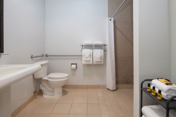 The image shows a clean, modern bathroom with a toilet, sink, shower with a curtain, towel racks, and toiletries on a stand with extra towels.