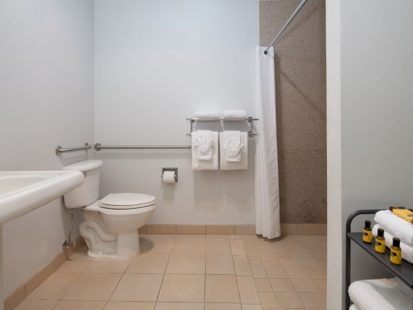 The image shows a clean, modern bathroom with a toilet, sink, shower with a curtain, towel racks, and toiletries on a stand with extra towels.