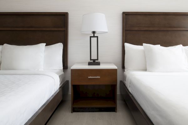 A hotel room setup with two neatly made beds featuring wooden headboards, a nightstand in between, and a lamp on top of the nightstand.