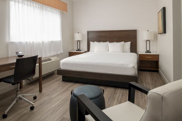 A modern hotel room with a bed, nightstands, lamps, a desk, chair, and window. The room has a minimalist design with neutral colors.