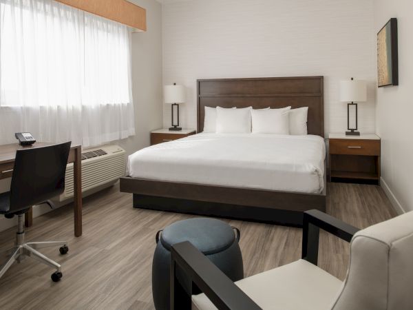 A modern hotel room with a bed, nightstands, lamps, a desk, chair, and window. The room has a minimalist design with neutral colors.