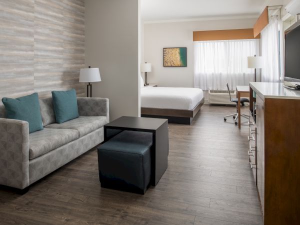 A modern hotel room features a sitting area with a sofa, a coffee table, and a bed in the background, along with a desk and a flat-screen TV.