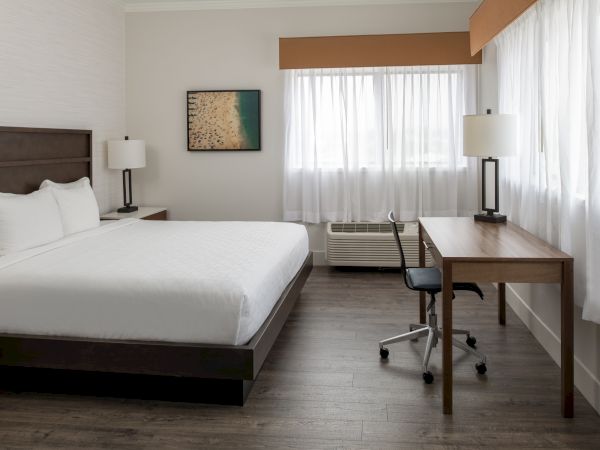 A tidy hotel room with a large bed, desk, chair, two lamps, and a window with sheer curtains. A beach image decorates the wall.