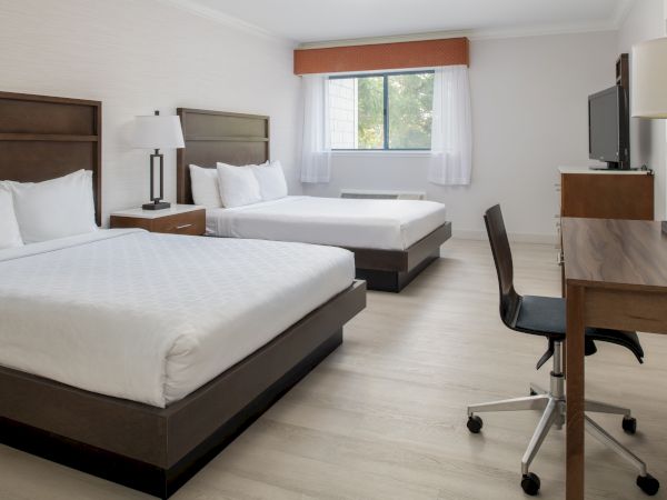 The image shows a clean, modern hotel room with two double beds, a desk with a chair, a TV, and a window with curtains.