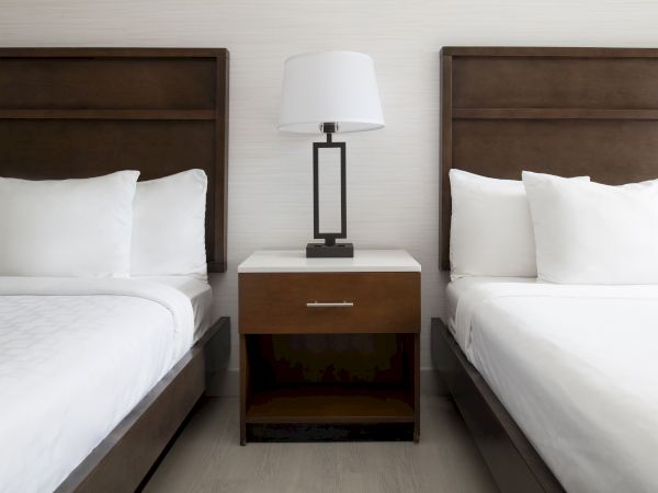 This image shows two neatly made beds with white linens and a wooden nightstand with a lamp in between them. The room is well-organized.