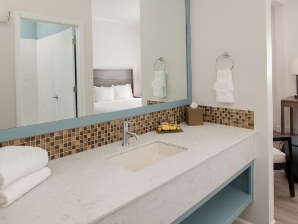 A modern bathroom with a white countertop, towels, a mirror, a sink, and a view into a bedroom and desk area in the background.