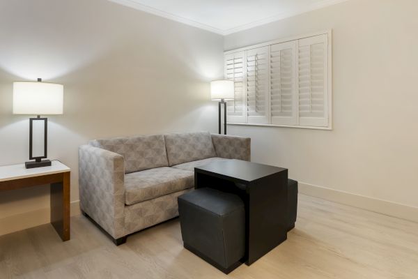 A modern, minimalist living room with a gray couch, two black ottomans, a black coffee table, two floor lamps, wooden table, and white shuttered windows.