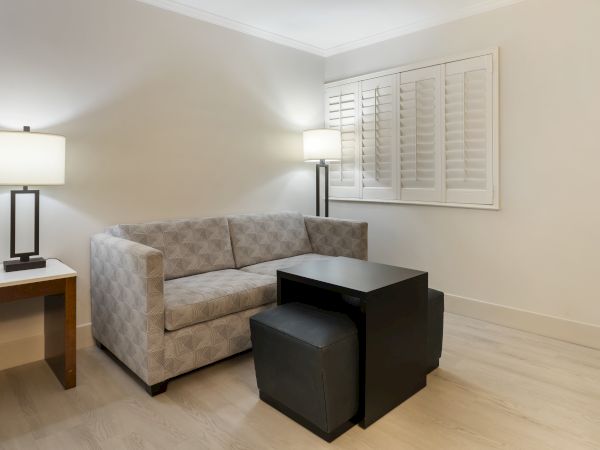A modern, minimalist living room with a gray couch, two black ottomans, a black coffee table, two floor lamps, wooden table, and white shuttered windows.