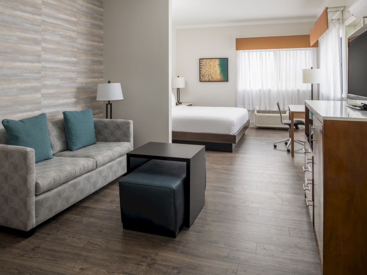 The image shows a modern hotel room with a sofa, two ottomans, a TV, a bed, a desk, and a chair in a spacious layout ending the sentence.