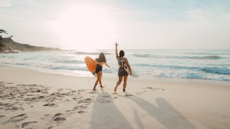 Two people with surfboards walk towards the ocean on a sandy beach as the sun sets, casting long shadows on the sand.