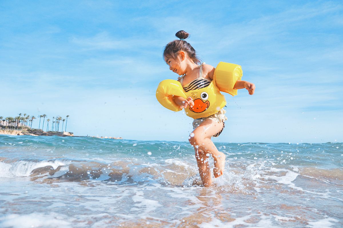 A young girl wearing inflatable arm floaties and a swimsuit is playing in the shallow waves of a beach under a blue sky with palm trees in the distance.