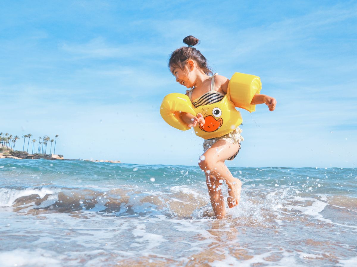 A young girl wearing inflatable arm floaties and a swimsuit is playing in the shallow waves of a beach under a blue sky with palm trees in the distance.
